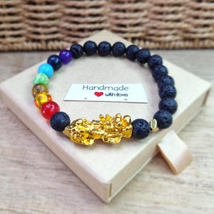 Puede incluir: Una pulsera de cuentas de piedra de lava negra porosa, una serie de cuentas de chakra multicolores y un dije central de dragón dorado. Incluye una etiqueta rectangular blanca con el texto Handmade with love y un corazón rojo.