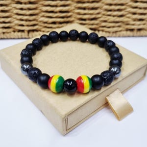 Può includere: Un braccialetto di perline con un motivo ripetuto di perline nere opache, perline di roccia lavica nera porosa e perline metalliche color argento. Il centro del braccialetto mostra due perline sferiche con strisce rosse, gialle e verdi.