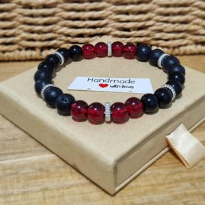 May include: A beaded bracelet featuring alternating black lava stone beads, smooth black beads, and translucent red beads. The bracelet includes silver-toned metal spacers and a white rectangular tag with the text Handmade with love and a red heart.
