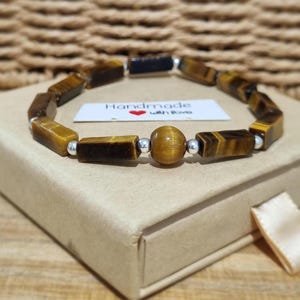 Könnte beinhalten: Ein Armband mit rechteckigen Tigerauge-Perlen und einer einzelnen runden Tigerauge-Perle, getrennt durch kleine silberfarbene Metallabstandshalter. Ein weißes Etikett mit dem Text "Handmade with love" und einem roten Herzsymbol ist zu sehen.