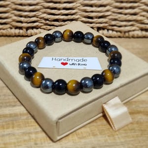 Könnte beinhalten: Ein Perlenarmband mit abwechselnd runden, polierten Tigerauge-, schwarzem Obsidian- und metallischen Hämatitperlen. Ein kleines weißes rechteckiges Etikett liegt in der Mitte mit dem Text Handmade with love und einem roten Herz.