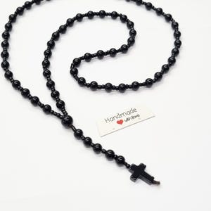 Könnte beinhalten: Eine lange Kette aus glänzenden schwarzen Perlen, die in einem Kreis angeordnet sind und an einem Ende ein kleines schwarzes Kreuz tragen. Ein weißes rechteckiges Etikett zeigt den gedruckten Text Handmade with love und ein kleines rotes Herzsymbol.