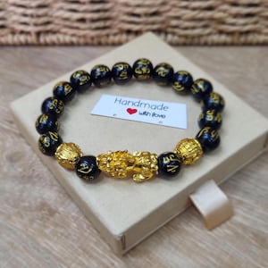 Könnte beinhalten: Ein Perlenarmband mit runden schwarzen Perlen mit goldfarbenen Inschriften, zwei strukturierten goldfarbenen Kugeln und einem zentralen goldfarbenen Anhänger in Form eines Fabelwesens. Ein weißes rechteckiges Etikett trägt die Aufschrift Handmade with love.