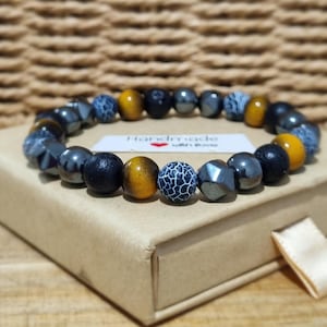 Pode incluir: Uma pulseira de contas com uma mistura de contas redondas e facetadas em preto, cinza metálico e amarelo dourado, juntamente com contas de padrão craquelado azul e branco. Uma pequena etiqueta branca com o texto Handmade with love está por baixo.