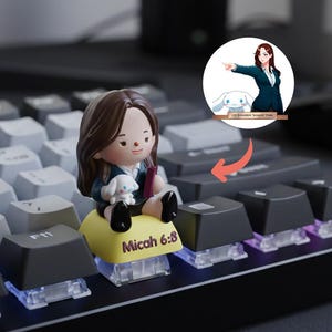 Puede incluir: Una tecla de teclado personalizada con una figura de una persona sosteniendo un perro blanco peque&ntilde;o y un libro, sentada en una tecla amarilla con el texto "Micah 6:8". Una ilustraci&oacute;n de una persona y un perro de dibujos animados est&aacute; en el fondo.