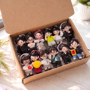 May include: A cardboard box filled with fourteen small, cartoon-style character keychains. Each keychain features a unique figure with distinct hair, clothing, and accessories, including some holding small items like flags or signs, arranged in a grid.