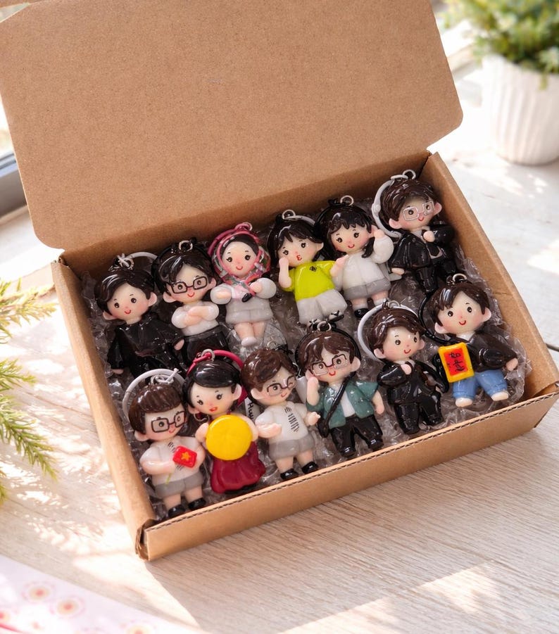May include: A cardboard box filled with fourteen small, cartoon-style character keychains. Each keychain features a unique figure with distinct hair, clothing, and accessories, including some holding small items like flags or signs, arranged in a grid.