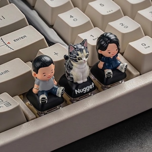 Puede incluir: Un teclado mec&aacute;nico con teclas personalizadas. Tres teclas presentan peque&ntilde;as figuras: un ni&ntilde;o con una camisa gris, un gato calic&oacute; y una ni&ntilde;a con un vestido azul. La tecla del gato tiene la palabra "Nugget". El teclado tiene teclas de color blanquecino.