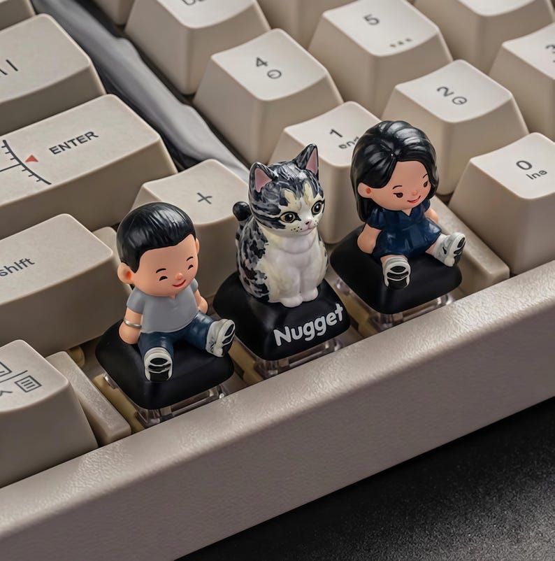 Puede incluir: Un teclado mec&aacute;nico con teclas personalizadas. Tres teclas presentan peque&ntilde;as figuras: un ni&ntilde;o con una camisa gris, un gato calic&oacute; y una ni&ntilde;a con un vestido azul. La tecla del gato tiene la palabra "Nugget". El teclado tiene teclas de color blanquecino.