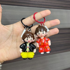 May include: Two character keychains featuring stylized figures with brown hair. One figure wears a black jacket over a light shirt with yellow pants on a black ring. The other figure wears a red floral patterned outfit on a red ring.