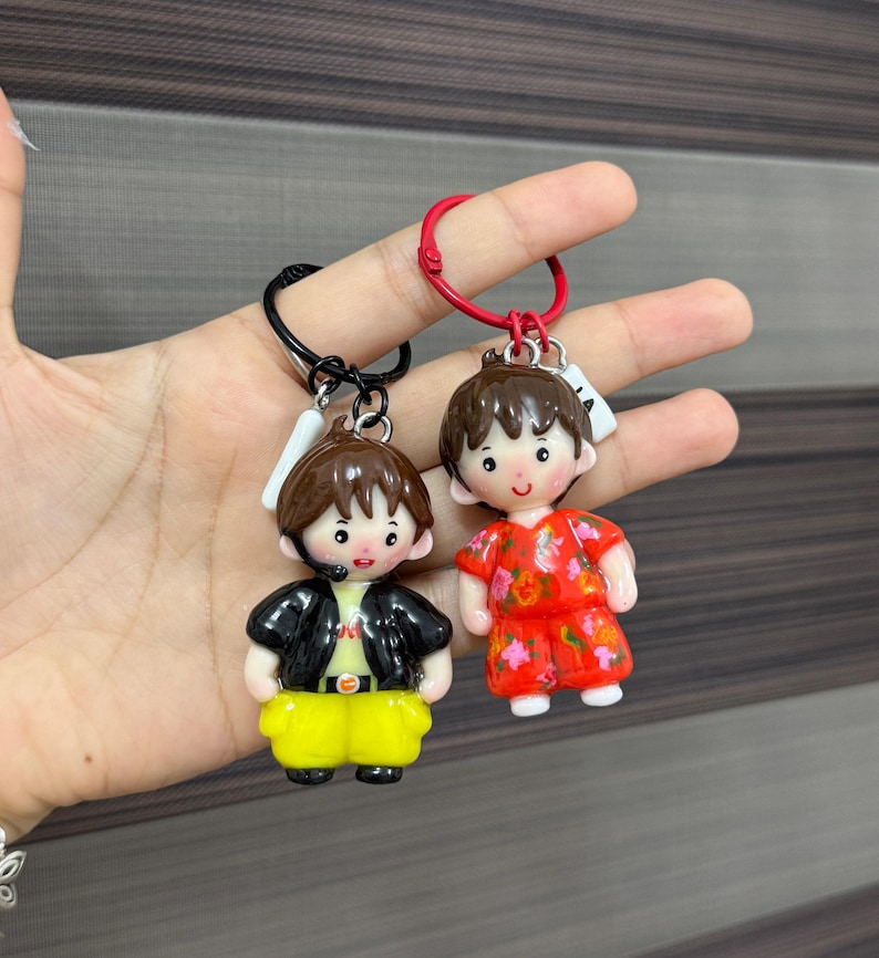 May include: Two character keychains featuring stylized figures with brown hair. One figure wears a black jacket over a light shirt with yellow pants on a black ring. The other figure wears a red floral patterned outfit on a red ring.