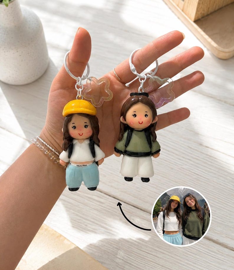 May include: Two custom character keychains featuring miniature figures with brown hair, attached to white metal rings with iridescent star charms. One figure wears a yellow cap, white top, and light blue pants; the other wears a green top and white pants.