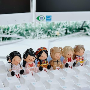 Puede incluir: Seis peque&ntilde;as y coloridas figuras de ni&ntilde;as sentadas con varios atuendos y peinados. Est&aacute;n dispuestas sobre las teclas blancas de un teclado. Las figuras tienen detalles y est&aacute;n en una variedad de colores, incluyendo rosa, rojo y marr&oacute;n.