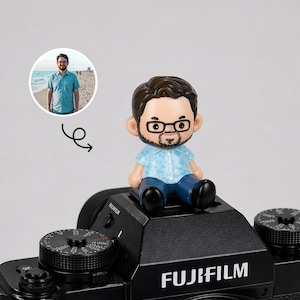 Könnte beinhalten: Eine kleine Figur eines Mannes mit Brille und Bart, gekleidet in einem hellblauen Hemd und blauen Hosen, sitzt auf einer schwarzen Fujifilm-Kamera. Ein kleines Foto eines Mannes in einem blauen Hemd befindet sich in der oberen linken Ecke. Auf der Kamera steht das Wort "FUJIFILM".