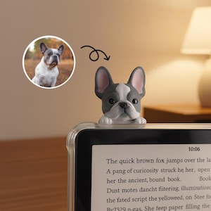 Op de afbeelding: Een grijze en witte Franse Bulldog-figuur zit bovenop een e-reader. Het beeldje heeft een witte vlek op het voorhoofd en roze oren. De e-reader toont tekst en de tijd 10:06.