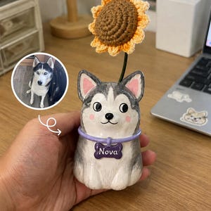 May include: A ceramic husky figurine with a purple collar and a bone-shaped tag reading Nova. The figurine features a gray and white coat, pink ears, and a crocheted sunflower attached to the top. A circular inset shows a photo of a real husky.