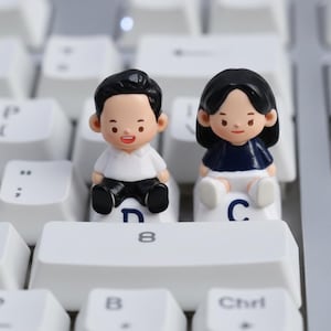 Puede incluir: Dos peque&ntilde;as figuras de un hombre y una mujer sentados en teclas de teclado blancas. El hombre viste camisa blanca y pantal&oacute;n negro. La mujer lleva top azul marino y shorts blancos. Las teclas del teclado son blancas.