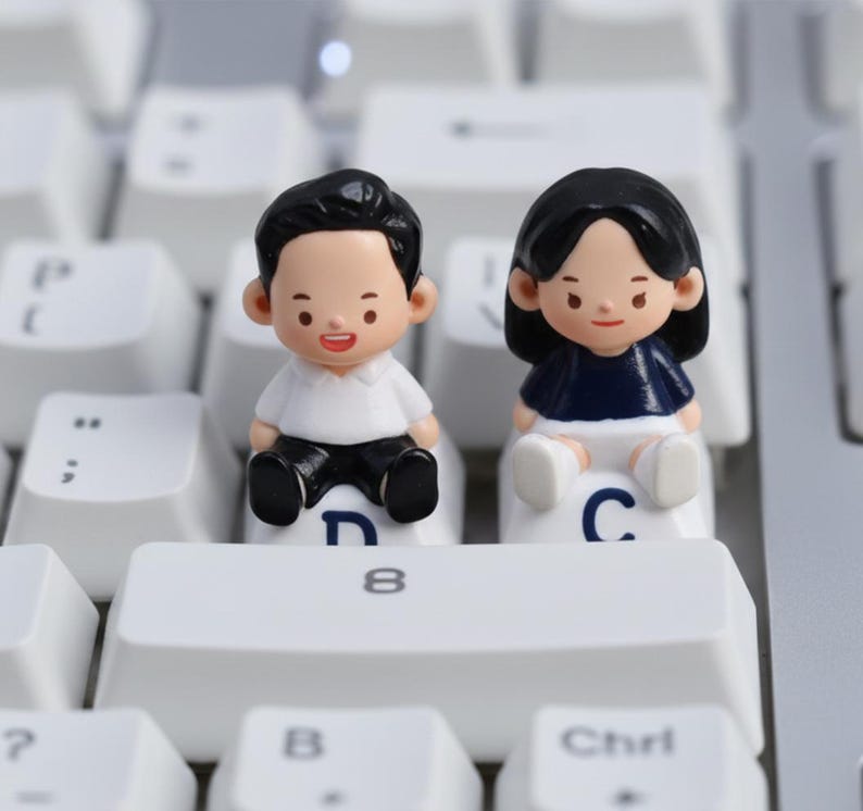 Puede incluir: Dos peque&ntilde;as figuras de un hombre y una mujer sentados en teclas de teclado blancas. El hombre viste camisa blanca y pantal&oacute;n negro. La mujer lleva top azul marino y shorts blancos. Las teclas del teclado son blancas.