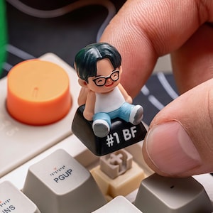 Puede incluir: Una peque&ntilde;a figura de una persona con gafas, camiseta blanca y pantalones azules, sentada en una tecla negra del teclado. La tecla est&aacute; etiquetada como "#1 BF". En el fondo se ve una tecla naranja.
