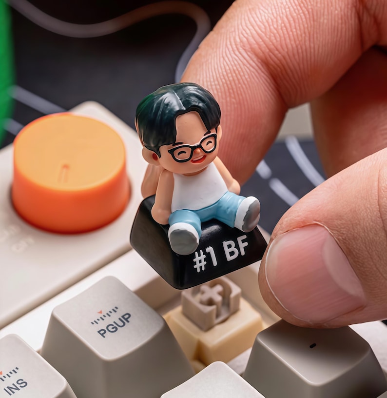 Puede incluir: Una peque&ntilde;a figura de una persona con gafas, camiseta blanca y pantalones azules, sentada en una tecla negra del teclado. La tecla est&aacute; etiquetada como "#1 BF". En el fondo se ve una tecla naranja.