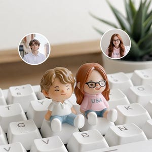 May include: Two miniature figurines of a boy and a girl sitting on a white keyboard. The boy figurine has light brown hair, a white shirt, and blue jeans. The girl figurine has red hair, glasses, a pink sweatshirt with "USA" on it, and blue shorts.
