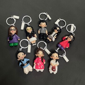 May include: A collection of eight individual keychains featuring small, hand-painted character figurines. Each figure has distinct hair, clothing, and accessories, including glasses, bags, and patterned outfits, all attached to white metal rings.