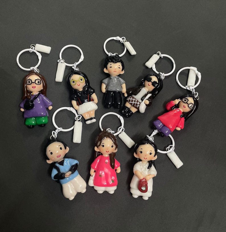May include: A collection of eight individual keychains featuring small, hand-painted character figurines. Each figure has distinct hair, clothing, and accessories, including glasses, bags, and patterned outfits, all attached to white metal rings.