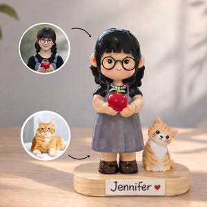 May include: A figurine of a girl with black hair and glasses holding a red apple, accompanied by a tabby cat. The base is wooden and has the name "Jennifer" and a heart symbol.