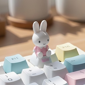 Peut inclure: Une figurine de lapin blanc vêtue d'une robe rose est posée sur une touche de clavier. Le clavier comporte des touches de différentes couleurs pastel, dont le bleu, le jaune et le rose. La touche sur laquelle le lapin est assis indique "Min".