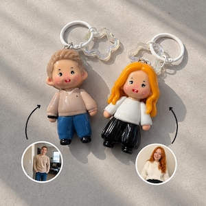 Puede incluir: Dos llaveros de personajes personalizados con figuras en miniatura y anillas blancas. Una figura tiene el pelo castaño corto, un suéter beige y pantalones azules. La otra tiene el pelo largo, ondulado y naranja, un suéter blanco y pantalones negros.