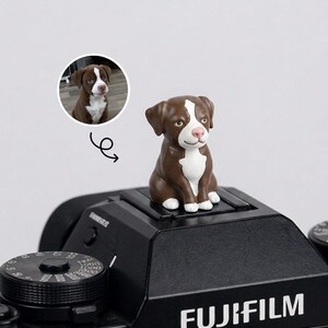 May include: A brown and white dog figurine sits on a black Fujifilm camera. The figurine has a white chest and face markings. A small circular image of the dog is in the upper left corner. The camera has a dial and the word "FUJIFILM" printed on it.