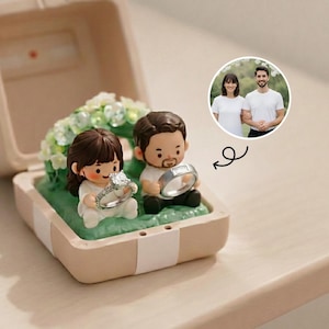 Könnte beinhalten: Eine beige Ringbox mit einer Miniatur-Paarfigur, die Ringe hält. Die weibliche Figur hält einen großen Diamantring, während die männliche Figur einen silbernen Ring hält. Im Hintergrund befindet sich ein kleines Foto eines Paares.