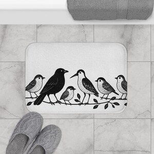 May include: White rectangular bath mat featuring a black and white illustration of six birds perched on a leafy branch. The birds vary in size and design, with a large black bird in the center. The mat is placed on a tiled floor.