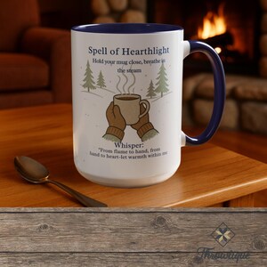 May include: White ceramic mug with a blue interior and handle. The mug features the text "Spell of Hearthlight" and an illustration of a steaming mug held by gloved hands, with pine trees. A spoon rests on the wooden table.