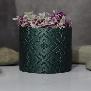 May include: A dark green cylindrical planter with a repeating floral pattern. The planter is filled with purple and green flowers. Several smooth, gray stones are arranged around the planter on a white surface.