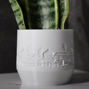 Könnte beinhalten: Ein weißer Keramik-Pflanztopf mit einem detaillierten Relief einer Stadtlandschaft. Der Topf enthält eine grüne Pflanze mit langen, gestreiften Blättern. Das Design zeigt Gebäude, Schornsteine und andere architektonische Elemente.