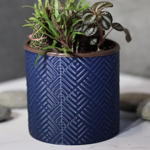 May include: A navy blue cylindrical planter with a geometric pattern, filled with a variety of small green and burgundy plants. The rim of the planter is brown. Several gray stones are scattered around the base.