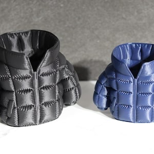 May include: Two miniature puffer jackets, one black and one blue, are displayed. Each jacket has a hood, a zipper, and a quilted design. The jackets are on a white surface against a gray background.