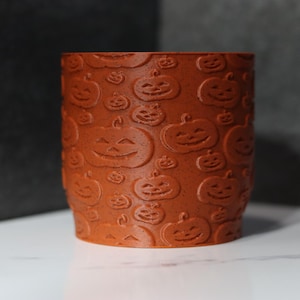May include: A cylindrical, orange ceramic container with a repeating pattern of carved jack-o'-lantern faces. The container is likely for Halloween decor or storage. The background is a dark gray wall and a white surface.