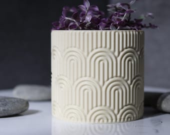 May include: A cream-colored, cylindrical planter with a repeating arch pattern, filled with trailing purple Wandering Jew plant. The planter is on a white surface with gray stones, creating a minimalist aesthetic. Ideal for home decor.