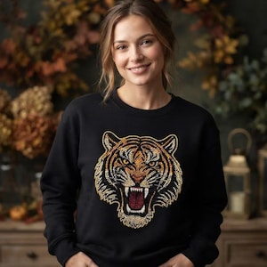 Può includere: Una felpa nera a maniche lunghe con una grande e dettagliata grafica di una testa di tigre sul petto. Il design della tigre è realizzato in tonalità arancione, giallo e bianco, con la bocca aperta che mostra i denti e una lingua rosso scuro.