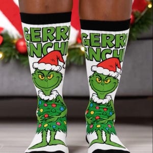 May include: White socks with a black cuff featuring a green Grinch character wearing a Santa hat and a Christmas tree-like outfit. The socks have the text "GERRINCH" in green letters.
