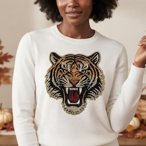 May include: A white long-sleeve sweater featuring a large, central tiger head graphic. The tiger design is composed of numerous small, shimmering beads in shades of orange, black, white, and red, depicting the animal with an open mouth.