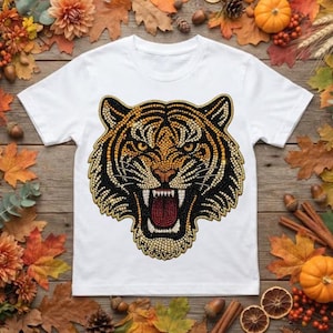 Puede incluir: Una camiseta blanca de manga corta con un gráfico grande y detallado de una cabeza de tigre. El diseño del tigre está compuesto por numerosas pequeñas piedras de estrás brillantes en tonos naranja, negro, blanco y amarillo, con la boca abierta.