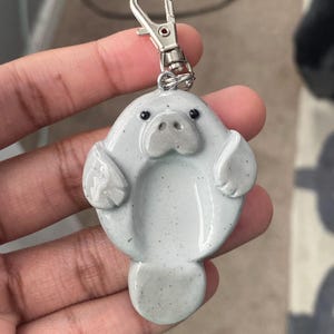 May include: A light grey manatee keyring with a silver clasp. The manatee has small, round eyes and a detailed face. The keyring is made of a smooth, glossy material.