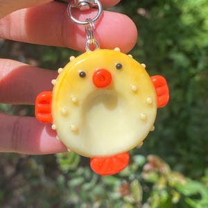 Op de afbeelding: Een gele en crèmekleurige kogelvis sleutelhanger met oranje vinnen en een rode neus. De sleutelhanger heeft een zilveren sluiting en ring. De kogelvis heeft kleine zwarte ogen en is versierd met kleine, ronde, crèmekleurige bultjes.