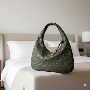 May include: Olive green woven leather shoulder bag with a curved design. The bag features a single handle and a decorative gold-toned accent. The bag is displayed on a neutral-toned ottoman in a bedroom setting.
