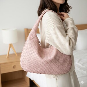 May include: A light pink woven shoulder bag with a curved shape. The bag has a long strap and a zipper closure. The woven pattern is visible across the entire surface of the bag. The bag is being held over the shoulder.
