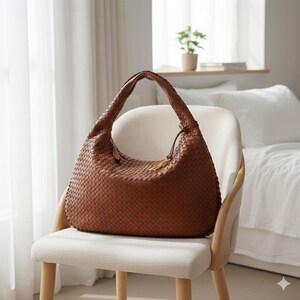 May include: A brown woven leather hobo bag with a single shoulder strap. The bag is sitting on a cream-colored chair with wooden legs. A gold-colored charm hangs from the zipper. The background includes a window and a bed.