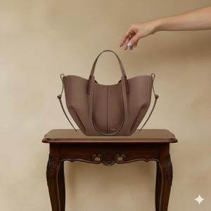 May include: A taupe-colored tote bag with a structured design, featuring a top handle and a detachable shoulder strap. The bag is placed on a dark brown wooden table with ornate carvings. The bag has a minimalist aesthetic.